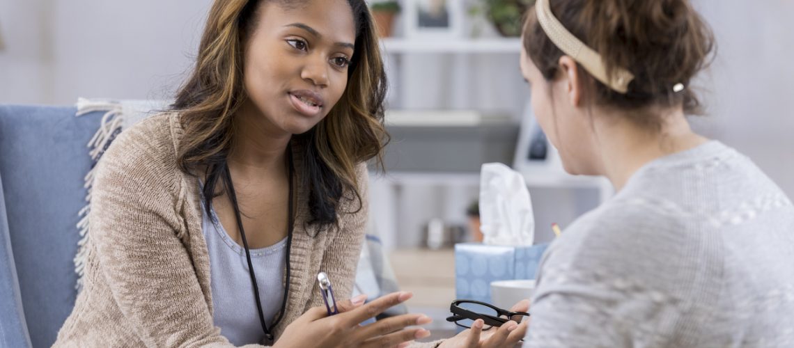 Serious female counselor gestures while talking with Caucasian female client. The counselor is holding eyeglasses and a pen. They are discussing serious issues.